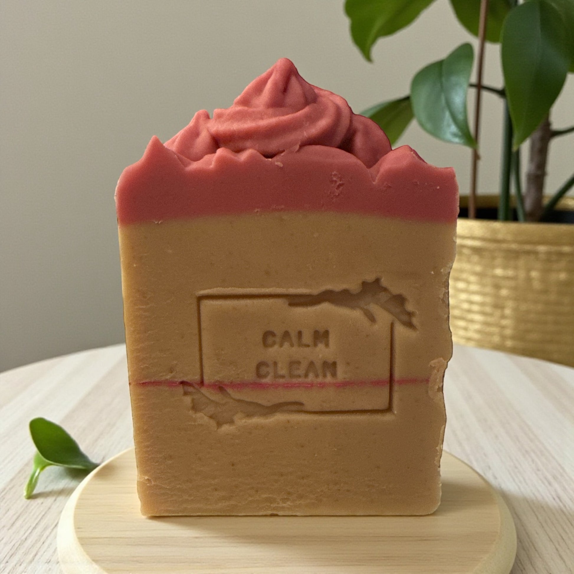 Handcrafted rectangular soap bar standing upright on a light wooden dish. The soap is tan with a thin pink line across the middle and a textured pink top. The front is stamped with the words “CALM CLEAN.”